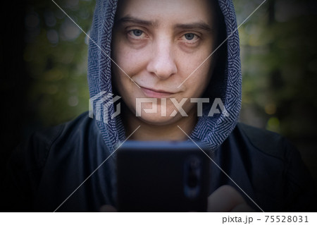 Concept. A man in a leather jacket with a hood looks at the phone and smiles like a maniac or a telephone scammer. Theft of information from a smartphone. Correspondence of a maniac on social networks Concept. A man in a leather jacket with a hood looks at the phone and smiles like a maniac or a telephone scammer. Theft of information from a smartphone. Correspondence of a maniac on social networks 75528031