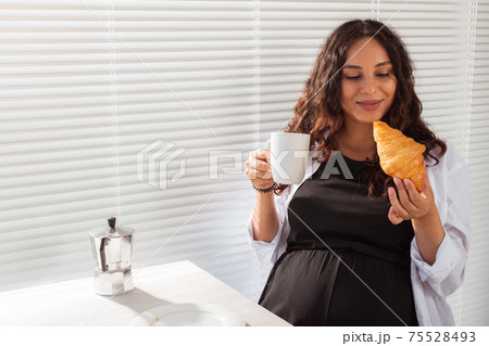 Happy pregnant young beautiful woman eating croissant during morning breakfast. Concept of pleasant morning and positive attitude during pregnancy 75528493