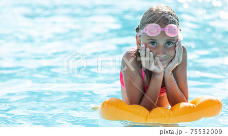 Young girl rests on floating pillow inside the pool with swimming goggles on her forehead, smiling 75532009