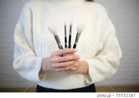 The girl holds brushes in her hands for makeup. In a white sweater, white brushes, on a black background. Soft selective focus. The girl holds brushes in her hands for makeup. In a white sweater, white brushes, on a black background. Soft selective focus. 75532167