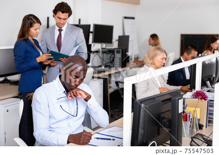 Puzzled african businessman sitting at desk in open plan office, thinking about business strategy 75547503