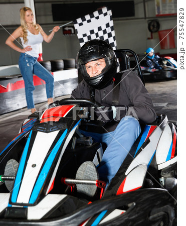 Man in helmet driving car for karting in sport club, woman with flag on background 75547829