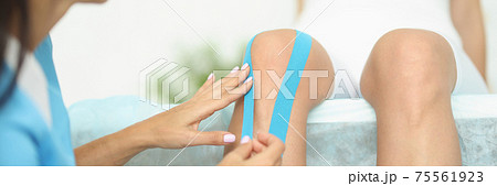 Doctor glues blue elastic strips to patient knee. Doctor glues blue elastic strips to patient knee. 75561923