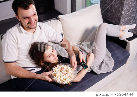 Young couple eating popcorn while watching movie lying on sofa 75564444