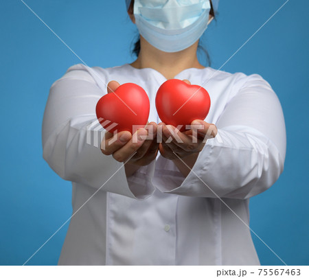 medic in a white coat, a mask stands and holds a red heart on a blue background medic in a white coat, a mask stands and holds a red heart on a blue background 75567463