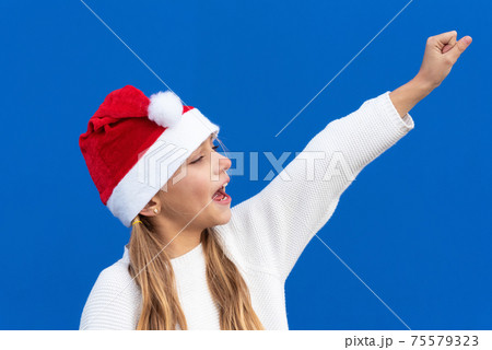 Joyful Young girl in Santa hat gesturing on blue background. Joyful Young girl in Santa hat gesturing on blue background. 75579323