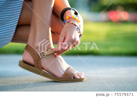 Close up of woman hands tying her open summer sandals shoes on sidewalk in sunny weather. 75579371