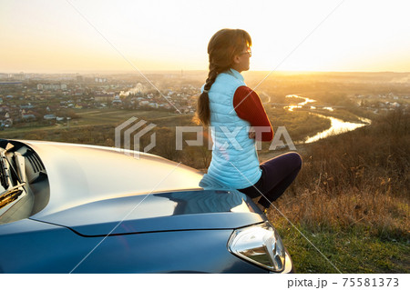 Young woman standing near her car enjoying warm sunset view. Girl traveler leaning on vehicle hood looking at evening horizon. Young woman standing near her car enjoying warm sunset view. Girl traveler leaning on vehicle hood looking at evening horizon. 75581373