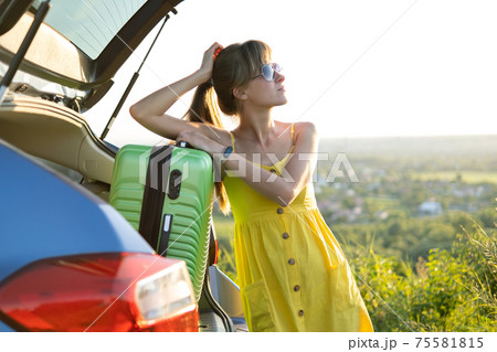 Young female driver having rest leaning on a suitcase in her car in summer field. Travel and vacations concept. 75581815