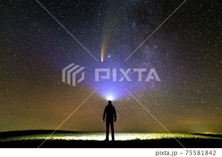Silhouette of a man with flashlight on his head pointing bright beam of light on starry sky with Neowise comet with light tail. 75581842
