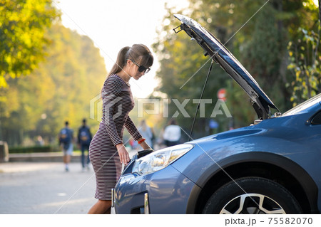 Puzzled female driver standing on a city street near her car with popped up hood looking at broken engine. Puzzled female driver standing on a city street near her car with popped up hood looking at broken engine. 75582070