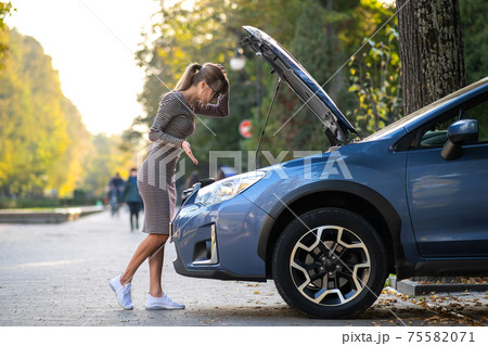 Puzzled female driver standing on a city street near her car with popped up hood looking at broken engine. 75582071