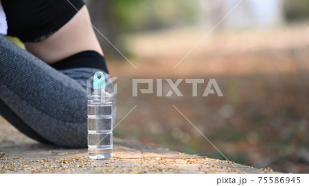 A bottle of water and sportswoman sitting and resting after workout at outdoor. 75586945