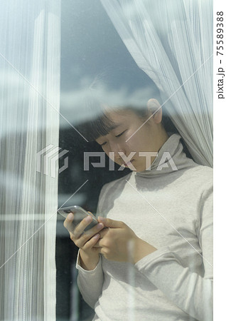 窓辺でスマートフォンを見る女の子（外出自粛要請のイメージ） 75589388
