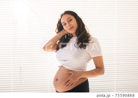 Smiling young beautiful pregnant woman touching her belly and rejoicing. Concept of positive and pleasant feelings while waiting for the baby 75590868