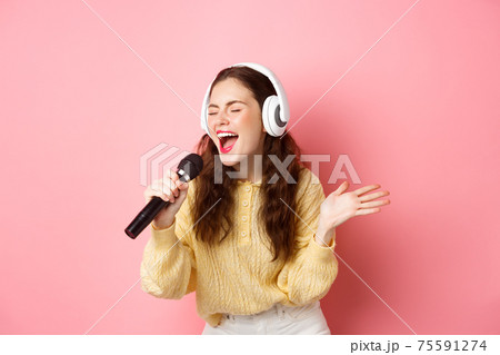Portrait of young woman singing karaoke, wearing headphones and performing song, holding microphone, standing against pink background 75591274