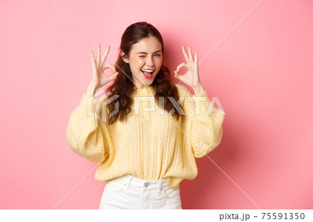 Portrait of cheerful pretty girl winking and saying yes, showing okay sign, alright gesture, praise good job, well done, standing pleased against pink background Portrait of cheerful pretty girl winking and saying yes, showing okay sign, alright gesture, praise good job, well done, standing pleased against pink background 75591350