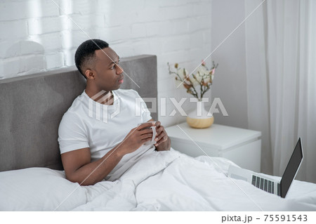 A young man in white tshirt feeling comfortable and having coffee in bed 75591543