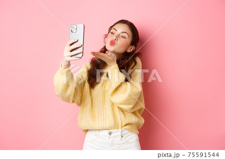 Beautiful flirty girl taking selfie on smartphone, sending air kiss during video chat, standing against pink background Beautiful flirty girl taking selfie on smartphone, sending air kiss during video chat, standing against pink background 75591544