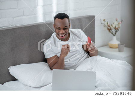A young man in white tshirt lying in bed and working remotely 75591549