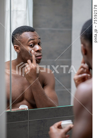 African american short-haired guy applying some cream on his face 75592578