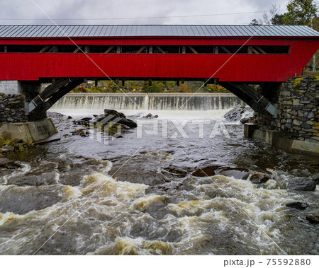 A red covered bridge first built in 1883 spans a rapidly flowing river with small hydroelectric A red covered bridge first built in 1883 spans a rapidly flowing river with small hydroelectric 75592880