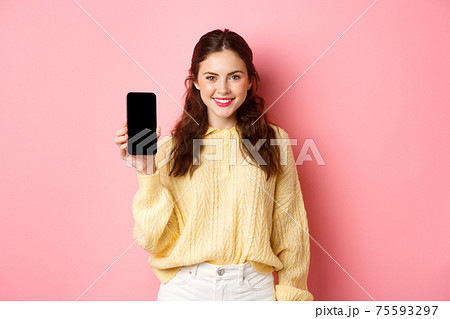 Technology and online shopping. Young smiling woman looking determined, advicing download app, showing smartphone app on screen, standing against pink background 75593297