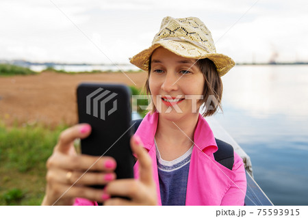 Young beautiful smiling woman is holding smart phone in hand and looking to the screen. Making selfie photo. Beach and a lake on a background. Young beautiful smiling woman is holding smart phone in hand and looking to the screen. Making selfie photo. Beach and a lake on a background. 75593915