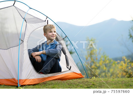 Happy child boy resting in a tourist tent at mountain campsite enjoying view of beautiful summer nature. Hiking and active way of life concept. Happy child boy resting in a tourist tent at mountain campsite enjoying view of beautiful summer nature. Hiking and active way of life concept. 75597660