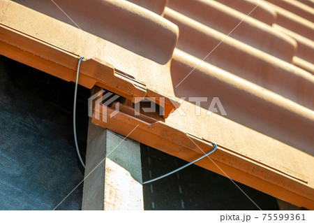 Closeup of metal montage anchor for installation of yellow ceramic roofing tiles mounted on wooden boards covering residential building roof under construction. Closeup of metal montage anchor for installation of yellow ceramic roofing tiles mounted on wooden boards covering residential building roof under construction. 75599361