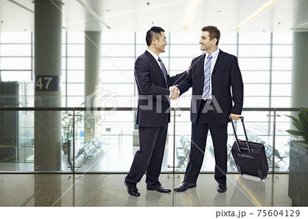 two businessmen, one asian and one caucasian, shaking hands and smiling at modern airport 75604129