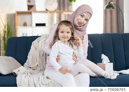 Pretty young Muslim mother and her cute little baby girl, sitting on the blue couch in modern appartment interior indoors. Mom cleans ear of her daughter with cotton swab. Close up 75606765