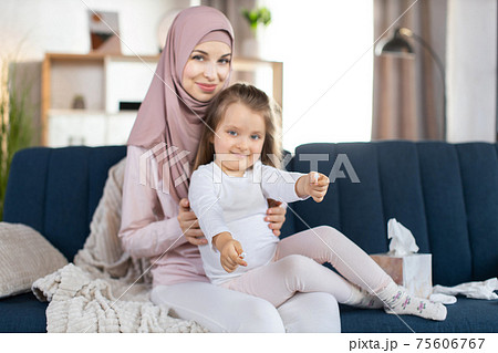 Young smiling Arabian mother with her cute little daughter, sitting on the blue sofa in cozy living room. Pretty child showing hygienic cotton swabs to camera. Hygiene and care concept 75606767