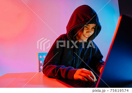 a contented girl in dark hooded clothes works at night at the computer. Hacker.  75619574