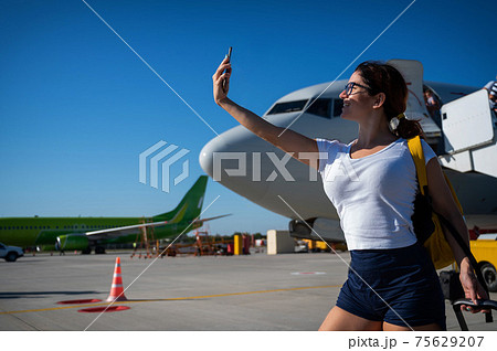 Caucasian red-haired woman taking a selfie on the background of the airplane. The girl is photographed on the runway before the flight 75629207