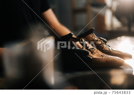 Close-up side view of hands of shoemaker shoemaker in black gloves holding old worn light brown leather shoes Close-up side view of hands of shoemaker shoemaker in black gloves holding old worn light brown leather shoes 75643833
