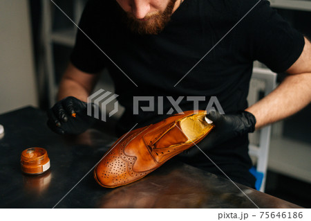 Close-up view of unrecognizable shoemaker in black gloves rubbing paint on light brown leather shoes with fingers. Close-up view of unrecognizable shoemaker in black gloves rubbing paint on light brown leather shoes with fingers. 75646186