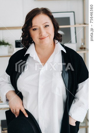 A businesswoman in a white shirt and trousers is standing with a hat in her hands standing in the office A businesswoman in a white shirt and trousers is standing with a hat in her hands standing in the office 75646360