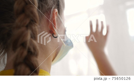 stay home coronavirus. kid looks out the window sad in a medical mask. concept pandemic coronavirus self-isolation infection stay home. kid mask touches glass covid 19 sunlight wants to go outdoors 75650126