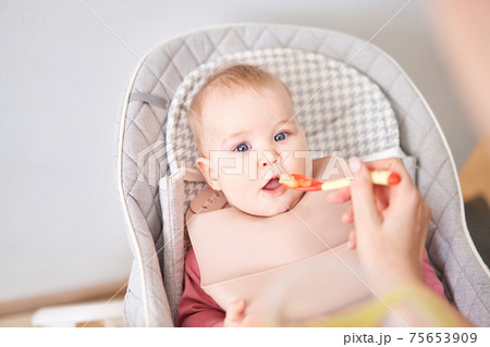 Young mother feeding her baby daughter with fruit puree. The baby is 7 months old, lying in a special high chair for babies. Young mother feeding her baby daughter with fruit puree. The baby is 7 months old, lying in a special high chair for babies. 75653909
