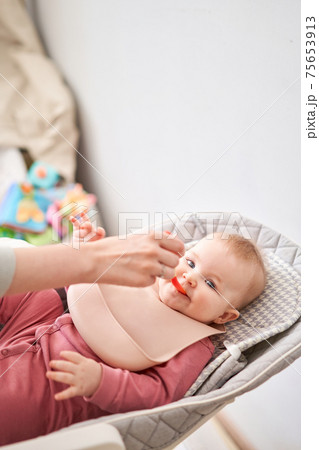 Young mother feeding her baby daughter with fruit puree. The baby is 7 months old, lying in a special high chair for babies. 75653913