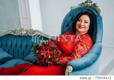 a woman in a red dress sits on a sofa and holds a bouquet of red roses and strawberries in the interior. a woman in a red dress sits on a sofa and holds a bouquet of red roses and strawberries in the interior. 75659412