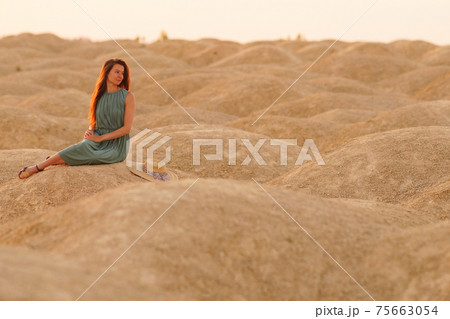 Young beautiful woman with long hair in blue dress sitting on sand at sunrise in desert. Looking thoughtful behind 75663054