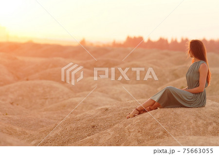Young beautiful woman with long hair in blue dress sitting elegant and thoughtful on sand at sunrise in sandy desert 75663055