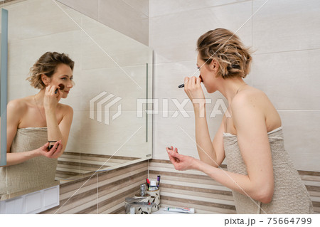 Woman applying blush while standing in front of the mirror in the bathroom Woman applying blush while standing in front of the mirror in the bathroom 75664799