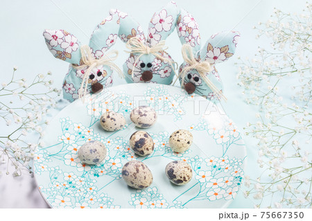 Easter composition. Three DIY textile Easter bunnies standing around plate with spring floral decor with quail eggs on pastel light blue. Horizontal orientation. Easter composition. Three DIY textile Easter bunnies standing around plate with spring floral decor with quail eggs on pastel light blue. Horizontal orientation. 75667350