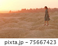 Young beautiful lonely smiling woman with long hair in long blue dress standing on sand at sunrise in hilly desert 75667423