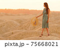 Young beautiful smiling woman with long hair in blue dress with straw hat in hand going on sand in desert at sunrise 75668021
