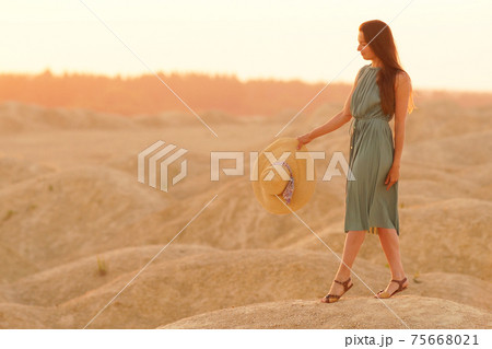 Young beautiful smiling woman with long hair in blue dress with straw hat in hand going on sand in desert at sunrise Young beautiful smiling woman with long hair in blue dress with straw hat in hand going on sand in desert at sunrise 75668021