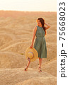 Young beautiful smiling woman with long hair in blue dress with straw hat in hand standing on sand in desert at sunrise 75668023
